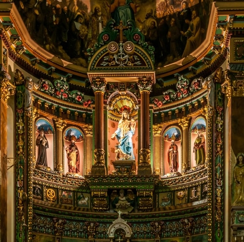 Altar de igreja ricamente decorado com uma estátua central da Virgem Maria vestida de azul e branco, ladeada por estátuas de santos em nichos dourados.